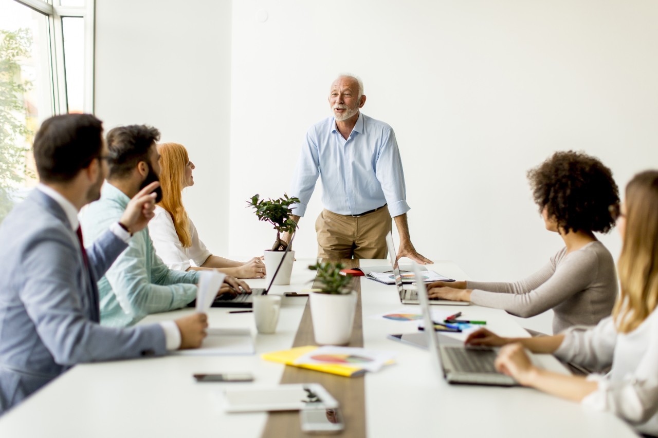 Senior businessman holds a meeting for younger colleagues, group, diverse, laptop, cup, conference room, man