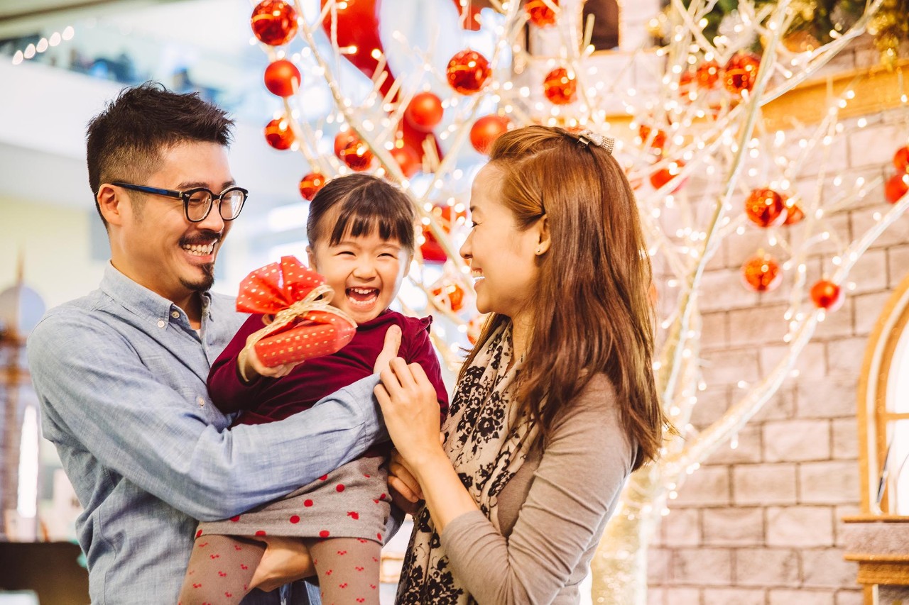 Young dad and young mom carrying little daughter who is holding a fancy wrapped Christmas gift while all smiling joyfully in front of the Christmas tree in a shopping mall, mother, father, child, girl, Asian
